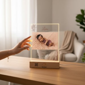 Può includere: Una cornice fotografica in acrilico illuminata personalizzata con base in legno. La cornice mostra una foto del bambino, il nome "Lara" e i dettagli della nascita. La cornice è illuminata con una luce calda e la base ha un codice QR e la parola "Memoria".