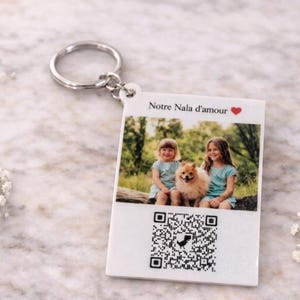 Peut inclure: Un porte-clés rectangulaire avec une photo de deux jeunes filles et un chien. Le porte-clés a un anneau et une chaîne argentés. Le texte "Notre Nala d'amour" est en haut, avec un cœur rouge. Un code QR est en bas.