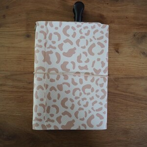 Puede incluir: Un cuaderno rectangular con una cubierta blanca y un patrón de leopardo en color tostado claro. El cuaderno se mantiene cerrado mediante una banda elástica fina de color claro envuelta horizontalmente alrededor del centro, con un clip negro arriba.