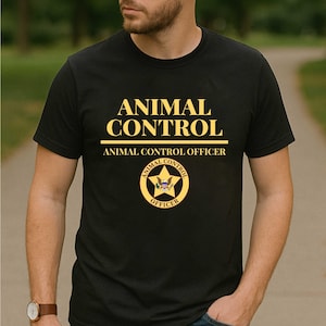 以下が含まれることがあります： 黒いTシャツ。前面には金色の文字で「ANIMAL CONTROL」と書かれ、その下に「ANIMAL CONTROL OFFICER」と書かれています。星と「ANIMAL CONTROL OFFICER」の文字が入った円形のバッジも表示されています。Tシャツが着用されています。