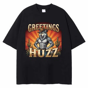 Op de afbeelding: Zwart t-shirt met een cartoonwolf met een cowboyhoed en spijkerbroek, met de tekst "GREETINGS HUZZ" in goud. De gespierde wolf staat voor een woestijnzonsondergang met cactussen.