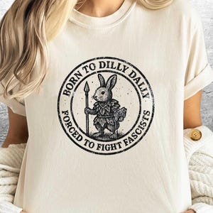 以下が含まれることがあります： クリーム色のTシャツ。円形のグラフィックがあり、槍と本を持ったウサギが描かれています。「BORN TO DILLY DALLY」と「FORCED TO FIGHT FASCISTS」の文字がかすれたフォントで書かれています。Tシャツは半袖です。