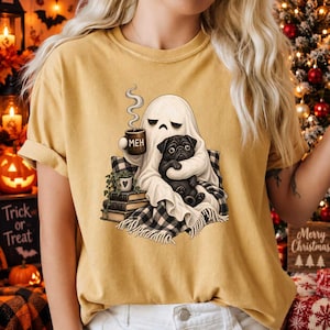 Może przedstawiać: Musztardowy t-shirt z grafiką przedstawiającą białego ducha trzymającego brązowy kubek z napisem 'MEH'. Duch siedzi na stosie książek, owinięty w czarno-biały koc w kratę i trzyma czarnego mopsa.