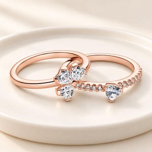 May include: Two rose gold rings featuring clear heart-shaped stones. One ring has a cluster of three heart-cut stones, while the other ring features a single heart-cut stone and a band accented with a row of small round clear stones.