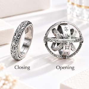 May include: A silver-coloured astronomical ring shown in two states: a closed band with intricate floral engravings and an expanded sphere featuring engraved zodiac symbols, text, and celestial markings. The text Closing and Opening appears below.