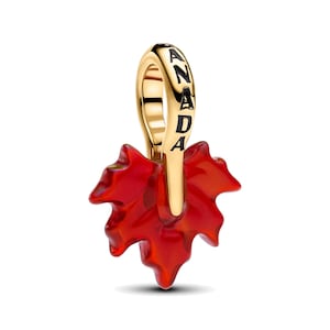 Può includere: Un ciondolo in metallo color oro a forma di foglia d'acero con una vivace finitura rossa traslucida. L'occhiello superiore presenta la parola CANADA incisa in lettere maiuscole nere, creando un netto contrasto con la superficie in metallo lucido.