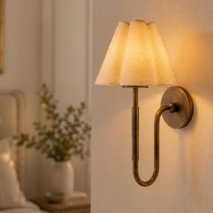 Op de afbeelding: Een wandlamp met een gebogen, antiek messingkleurige metalen arm en een ronde muurplaat. De lamp is voorzien van een geplooide, gebroken witte stoffen kap die een warm, zacht licht uitstraalt en de muur eromheen verlicht.