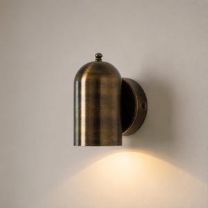May include: A wall-mounted light fixture with a cylindrical, dome-topped body finished in a brushed antique brass tone. The fixture features a circular base plate and emits a warm, downward-facing glow that illuminates the wall surface below.