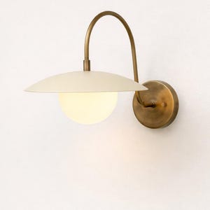 Könnte beinhalten: Wandleuchte mit cremefarbenem Schirm und kugelförmiger, leuchtender Glühbirne. Die Leuchte hat einen gebogenen, messingfarbenen Arm und einen kreisförmigen Sockel. Das Design ist minimalistisch und modern, geeignet für die Innenbeleuchtung.