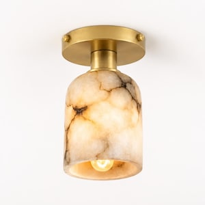 May include: A flush mount ceiling light fitting featuring a cylindrical shade made of translucent, cream-coloured stone with brown veining. The fitting includes a circular brass-toned metal canopy and a single exposed light bulb at the base.