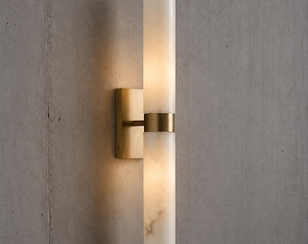 May include: A wall sconce featuring two vertical cylindrical white stone shades with a marbled texture, separated and held by a central brushed brass metal band and a rectangular brass wall plate, emitting a soft warm glow from within.