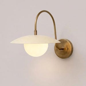 May include: A wall sconce featuring a circular brass mounting plate and a curved brass arm. The light fitting includes a wide, off-white saucer-shaped shade positioned above a spherical, frosted white glass globe that emits a soft, warm glow.