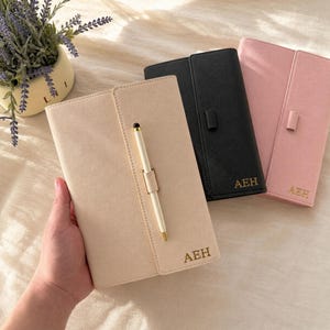 Könnte beinhalten: Drei rechteckige Notizbücher in Beige, Schwarz und Rosa, jeweils mit einer vertikalen Stifthalterung und einem weiß-goldenen Stift. In der unteren rechten Ecke jedes Notizbuchs sind die goldenen Buchstaben AEH eingeprägt. Die Einbände sind texturiert.