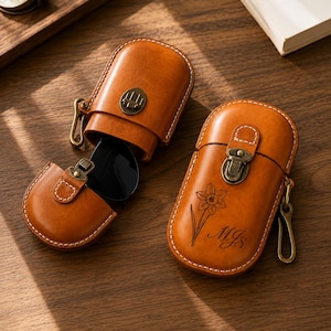 Könnte beinhalten: Zwei hellbraune Lederetuis für Brillen mit weißen Nähten und antikem Messingbeschlag. Ein Etui ist geöffnet und zeigt eine schwarze Sonnenbrille im Inneren. Das andere geschlossene Etui ist mit einer gravierten Narzisse und den Initialen MJs versehen.