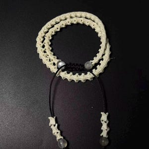May include: A double-strand bracelet constructed from off-white, segmented vertebrae-like bone beads. The strands are connected by a black cord featuring a sliding knot closure, two small translucent beads, and two dangling ends tipped with bone beads.