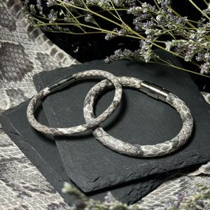 May include: Two circular bracelets featuring a textured, grey and white snakeskin pattern. Each bracelet is constructed with a smooth, cylindrical band and secured by a polished silver-toned metal magnetic clasp closure.