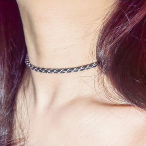Peut inclure: Un collier ras-du-cou tressé et fin, composé d'un cordon gris anthracite entrelacé d'un fil métallique argenté. Ce design crée un motif répétitif en forme de losanges qui entoure le cou dans un style ajusté et près de la peau.