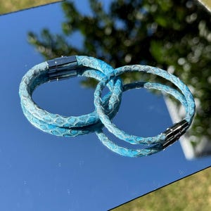 May include: Two circular bracelets featuring a textured, scale-like pattern in shades of bright blue and teal. Each bracelet is finished with a polished silver-toned cylindrical magnetic clasp that connects the two ends of the band securely.