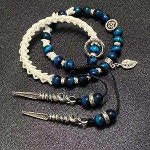 May include: A collection of beaded jewelry featuring blue and silver accents. The pieces include a white bone-like bracelet, a beaded necklace with a leaf charm, and two silver dagger-shaped pendants. The beads are a deep blue color.