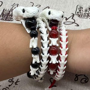 May include: Two bracelets featuring white snake head charms with black eyes. One bracelet has alternating black beads and white bone-like segments. The other bracelet features red beads and white bone-like segments on a red cord base.