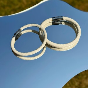 May include: Two off-white braided bracelets with a textured, scale-like pattern. Each bracelet features a cylindrical silver-toned metal magnetic clasp that connects the ends of the cord, creating a circular shape with a smooth, uniform finish.