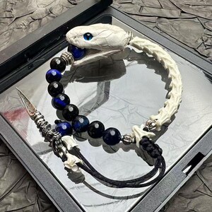 Puede incluir: Una pulsera que presenta una cabeza de serpiente blanca con ojos azules, una serie de cuentas esféricas de color azul oscuro y negro, y un segmento blanco similar a un hueso. Incluye un dije de daga plateado y cordones negros.