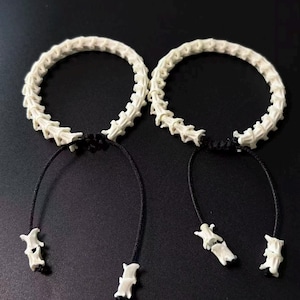 May include: Two bracelets made of off-white, segmented vertebrae bones arranged in a circular shape. Each bracelet features a black adjustable cord closure with two small, loose vertebrae bone beads hanging from the ends of the black strings.
