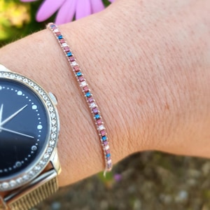 Peut inclure: Un bracelet délicat orné de petites perles rectangulaires roses, bleues et blanches. Le bracelet est porté sur un poignet, à côté d'une montre argentée avec un cadran noir et une lunette sertie de cristaux. L'arrière-plan est flou avec des touches de vert et de rose.