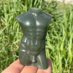 Peut inclure: Une sculpture de torse en pierre sculptée, vert foncé. La sculpture représente une forme masculine avec une musculature définie. La figurine est tenue dans une main sur fond d'herbe verte. La sculpture mesure environ 10 cm de haut.