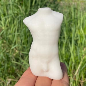 Peut inclure: Petite sculpture blanche représentant un torse masculin. La sculpture est lisse et détaillée, avec une musculature définie. La figurine est tenue dans une main sur fond d'herbe verte.