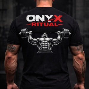 Peut inclure: T-shirt noir avec le texte "ONYX RITUAL" en blanc et rouge. Le t-shirt présente un graphique d'un haltérophile soulevant une barre. L'image est une vue arrière d'une personne avec des bras musclés et des tatouages.