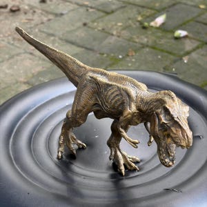 Puede incluir: Una figura de Tyrannosaurus rex de color bronce con un acabado de superficie texturizado y desgastado. El dinosaurio se muestra en una postura de pie, inclinado hacia adelante, con la boca ligeramente abierta y detalles de escamas.