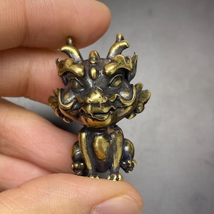 May include: A small, detailed brass figurine of a mythical creature. The figurine has a smiling face, horns, and ornate detailing. The creature is in a seated position, with its paws visible. The brass has a polished, golden appearance with darker accents.