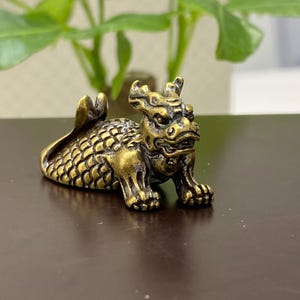 Puede incluir: Pequeña figura de latón de estilo antiguo que representa una criatura mítica, posiblemente un dragón. La figura tiene escamas detalladas, una expresión feroz y está en posición de reposo. El latón tiene una pátina cálida y envejecida.
