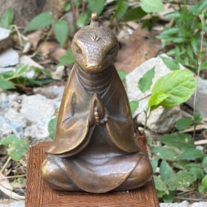 May include: A bronze-coloured figurine depicting a creature with a textured, snake-like head and closed eyes, wearing a robe with wide sleeves. The figure is in a seated meditation pose with hands pressed together in a prayer position holding beads.