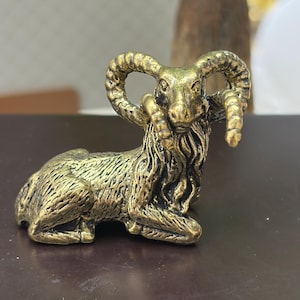 May include: A small, metallic gold-toned figurine of a ram in a resting position. The sculpture features detailed, textured fur, prominent curved horns, and a beard, with dark antiqued accents highlighting the intricate carved details.