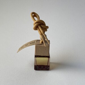 Op de afbeelding: Een klein, rechthoekig houten blok met een glazen flesje met een gele vloeistof. Een geknoopte, bruine veter is aan de bovenkant bevestigd. Een klein label met het woord "RUUM" is aan het blok bevestigd.