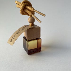 Può includere: Un deodorante per ambienti con un design di lacci annodati di colore marrone chiaro. Il deodorante ha un blocco di legno e una piccola bottiglia di vetro ambrato con la scritta "RUUM". È attaccata anche una piccola etichetta con la scritta "RUUM".