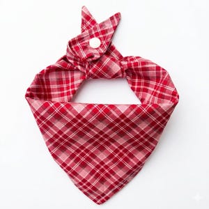 May include: Red and white plaid dog bandana with a button closure. The bandana is made of a soft fabric and features a classic plaid pattern in shades of red and white. The bandana is designed to be worn around a dog's neck.