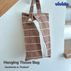 May include: A brown fabric hanging tissue bag featuring horizontal white dashed stripes, a single top handle, and a light-colored button closure. A white tissue is visible protruding from the center opening. The text vividopet is in the corner.