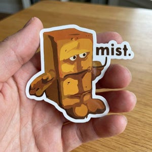 May include: A sticker featuring a cartoon character resembling a brick with arms and legs, in shades of brown and yellow. The character has a grumpy expression and the word "mist." is written in white text.