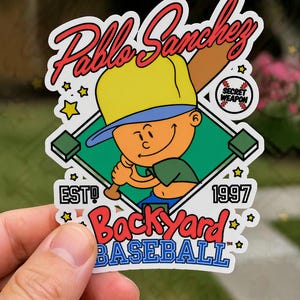 May include: A sticker featuring a cartoon boy in a yellow cap holding a baseball bat. The sticker has the name "Pablo Sanchez" in red script, with "Backyard Baseball" in red and blue text. The sticker also includes the text "EST! 1997" and a baseball-themed graphic.