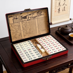 Pu&ograve; includere: Una custodia rettangolare rossa per set da mahjong con chiusura in metallo, contenente tessere color avorio con simboli neri, rossi e verdi. Il coperchio interno presenta un rivestimento in carta beige con illustrazioni e caratteri cinesi.