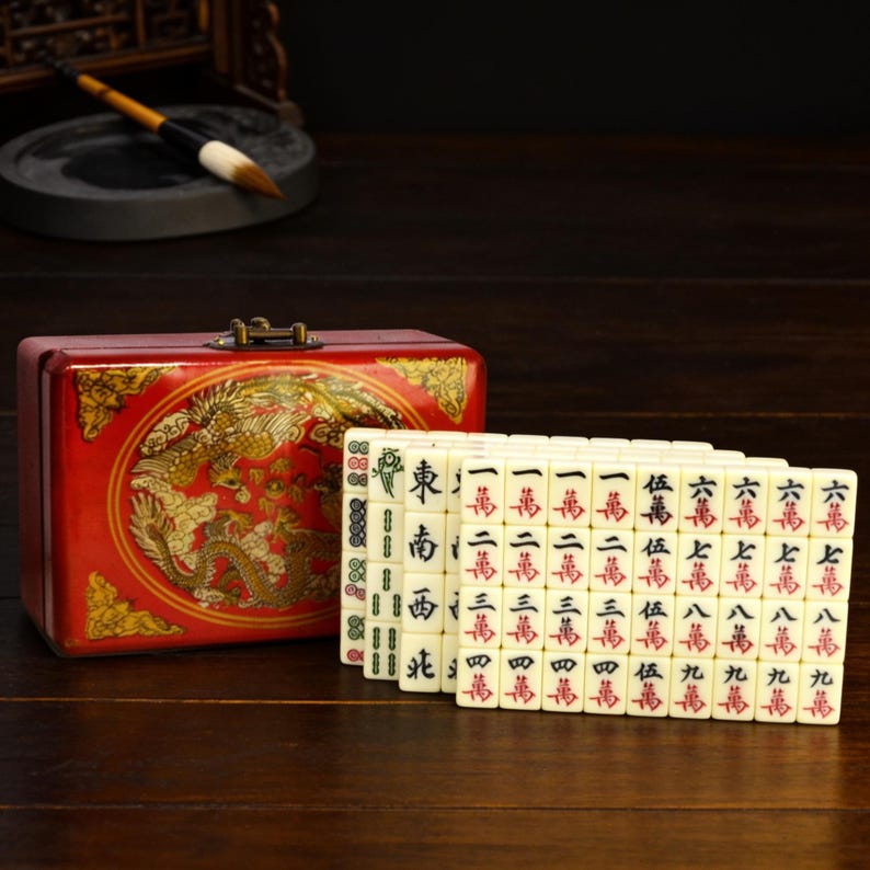Op de afbeelding: Een rode rechthoekige opbergdoos met een gouden drakenillustratie, vergezeld van een set gebroken witte Mahjong-tegels gerangschikt in rijen. De tegels tonen zwarte en rode Chinese karakters en symbolen, inclusief richtingsmarkeringen en cijfers.