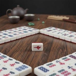 May include: A set of white rectangular Mahjong tiles with various red, blue, and green engraved symbols. One single tile is positioned upright in the centre, displaying a red character symbol, while other tiles are arranged in rows.