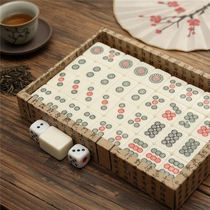 Pu&ograve; includere: Un set di tessere per mahjong color bianco sporco con motivi circolari verdi e rossi, disposte in una scatola di cartone rettangolare con calligrafia cinese nera. Due dadi bianchi e una tessera vuota sono posizionati fuori dalla scatola.
