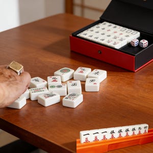 May include: A Mahjong set with white tiles featuring various symbols and characters. The set includes a red and black case, dice, and an orange tile rack. The tiles are scattered on a wooden surface.