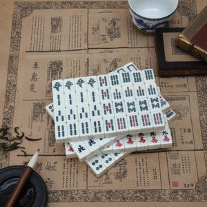 Pu&ograve; includere: Un set di tessere da mahjong bianche con caratteri e simboli incisi in nero, verde e rosso, disposte in una pila. Le tessere presentano vari contrassegni numerici e direzionali, inclusi i semi di bamb&ugrave;, caratteri e venti.