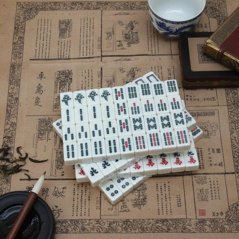 Pu&ograve; includere: Un set di tessere da mahjong bianche con caratteri e simboli incisi in nero, verde e rosso, disposte in una pila. Le tessere presentano vari contrassegni numerici e direzionali, inclusi i semi di bamb&ugrave;, caratteri e venti.