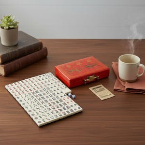 May include: A mahjong set featuring a rectangular red storage case with gold-toned metal latches, a collection of off-white tiles with black, red, and green character markings, two small blue and white dice, and a small rectangular instruction card.
